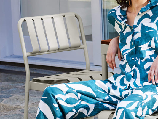 Woman in blue patterned outfit sitting on a beige metal outdoor chair, another chair in the background, on a stone patio.