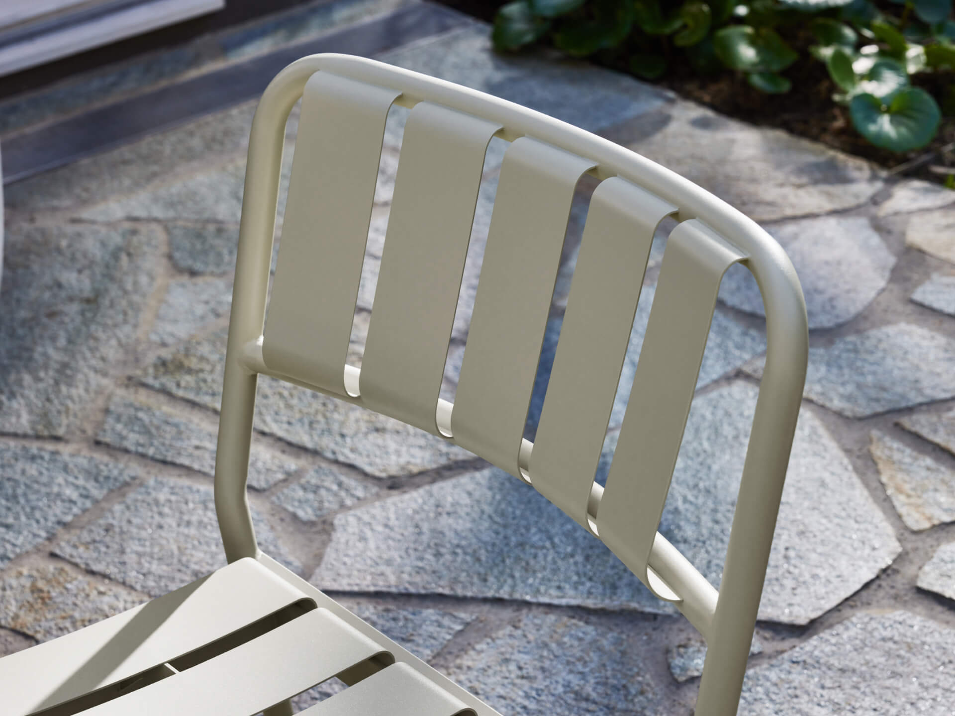 Beige metal outdoor chair with slatted backrest on a stone patio, surrounded by green plants in the sunlight.