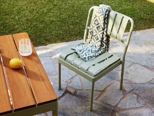 Outdoor setting with a light green metal chair draped with a patterned blanket, beside a wooden table with a striped glass and a lemon. Stone patio and grass.