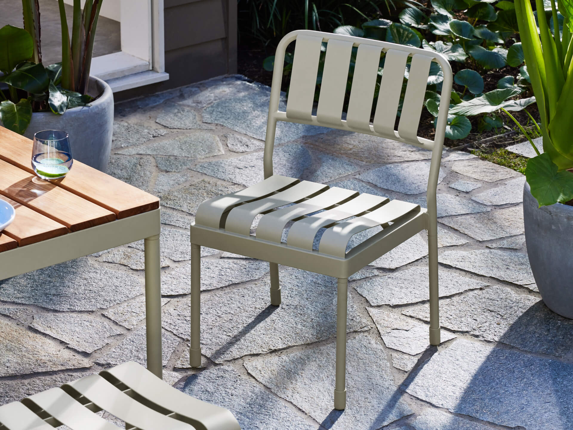 Outdoor setting with a light grey slatted metal chair on stone tiles, beside a wooden table. Potted plants surround the patio area.