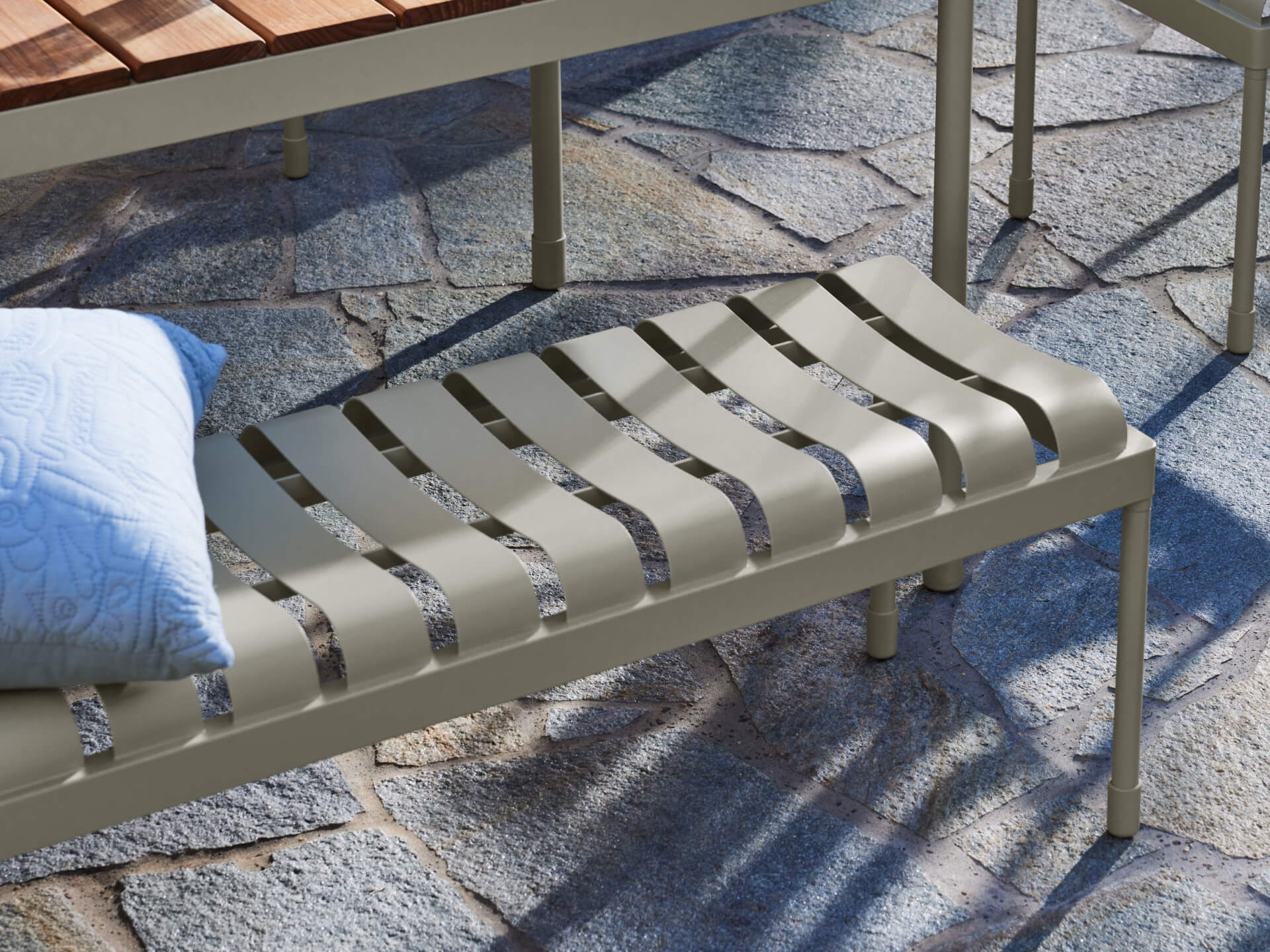 Metal outdoor bench with light green slats on stone patio, featuring a white pillow with blue floral pattern.