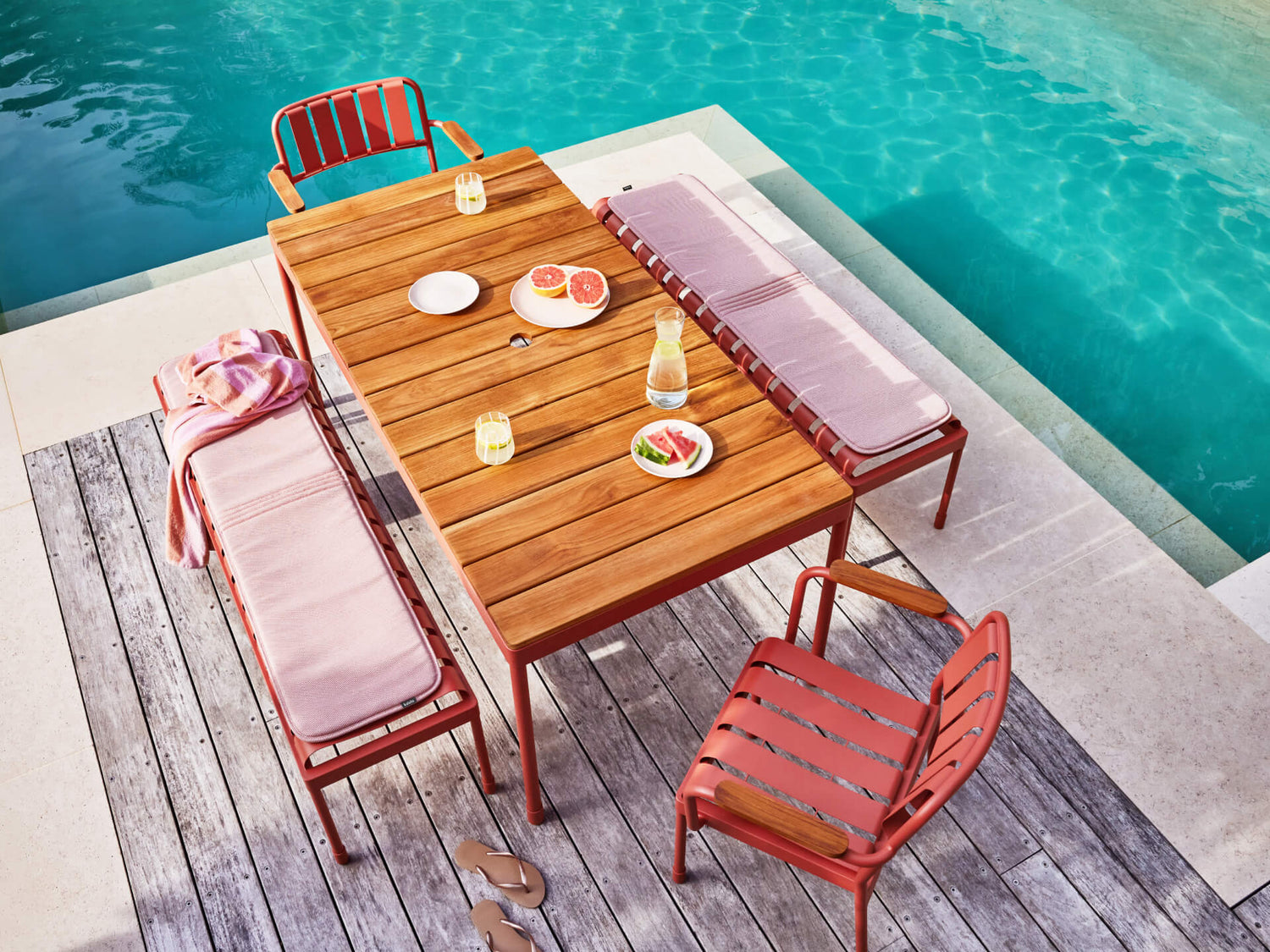 Outdoor wooden dining table with pink seating by a pool. Table set with drinks and fruit. Pink striped towel draped over one bench.