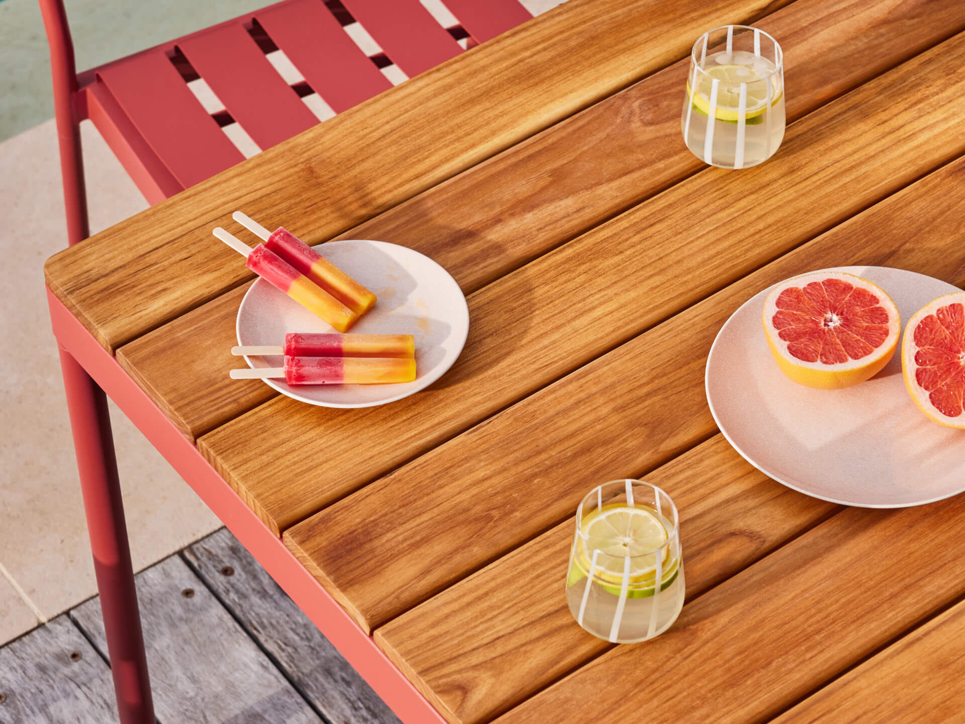 Wooden outdoor table with red metal chairs, holding plates of grapefruit and ice blocks, and glasses of lemon water.