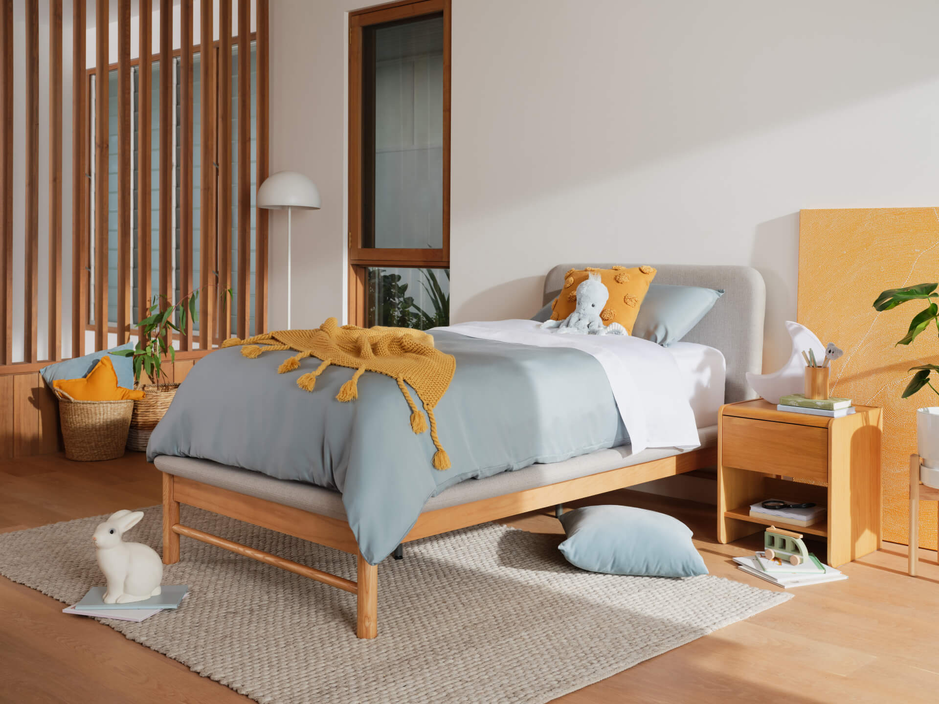 Modern bedroom with a light timber bed, grey bedding, mustard throw, and pillows. Wooden side table, indoor plants, and a woven rug.