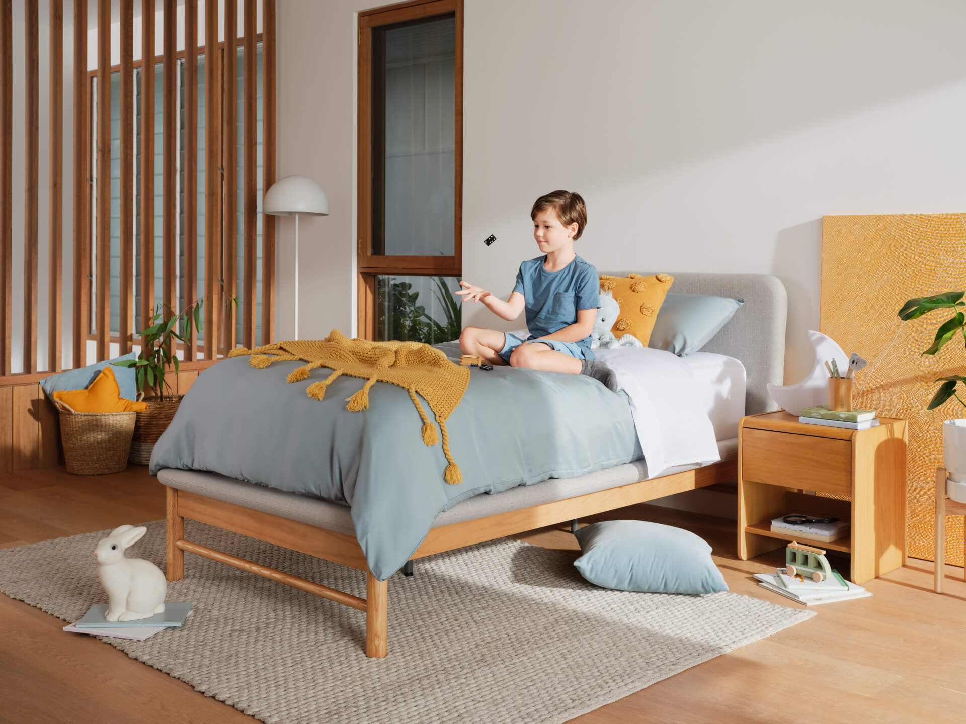 Child on a wooden bed with light grey bedding, mustard throw, and orange cushions. Room features a rug, plant, lamp, and decorative rabbit.