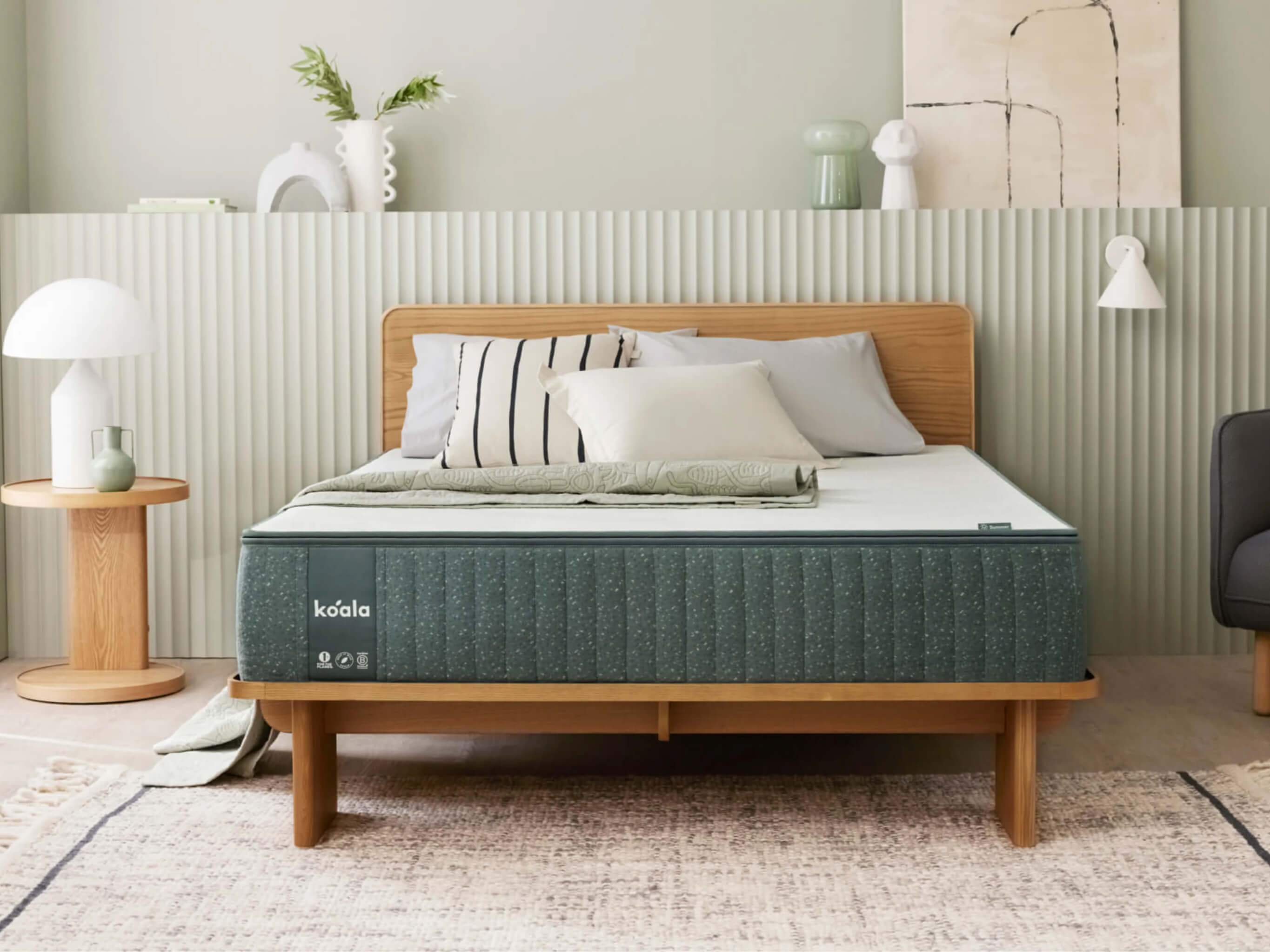 Minimalist bedroom with a wooden bedframe, green mattress, and white bed linens. A side table with a white lamp and decorative objects in the background.