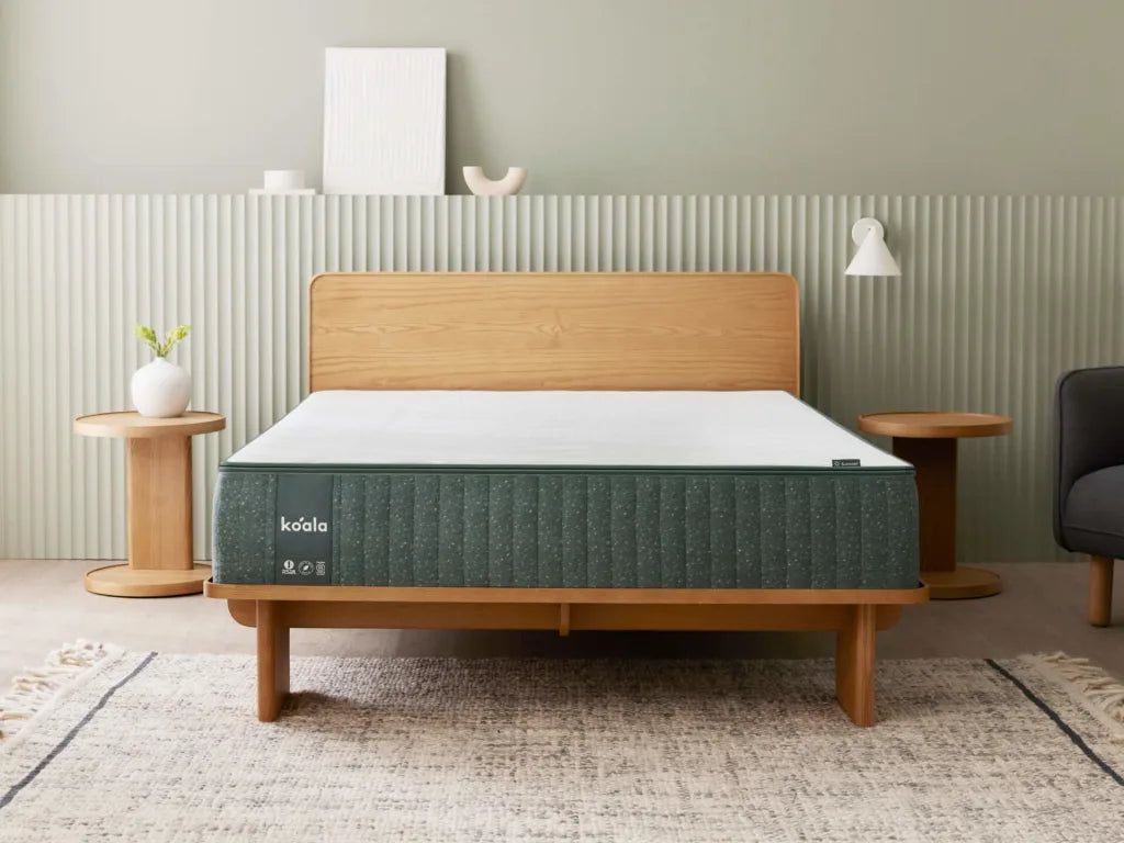 Bedroom with a wooden bed frame, green mattress, and white bedding. Flanked by round wooden side tables, set against a light green wall.