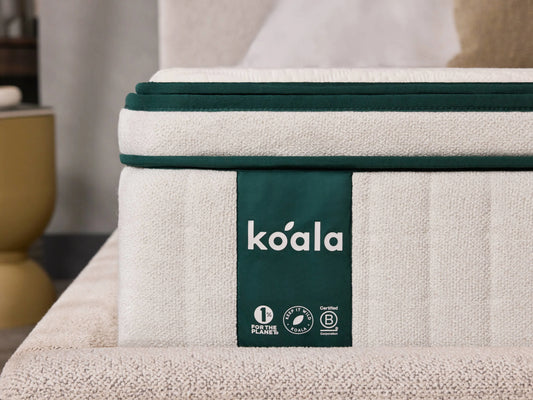 Close-up of a cream mattress with green trim, displaying a "koala" label and eco-friendly certifications in a modern bedroom setting.