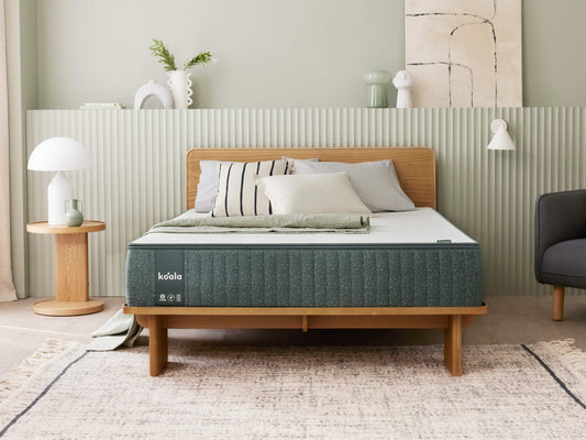 Modern bedroom with a wooden bed frame, dark green mattress, and striped pillows. Light grey walls, wooden side table, and decor accents.