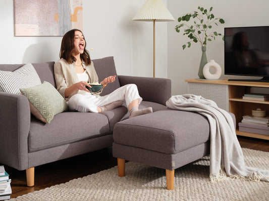 Woman relaxing on a grey sofa with matching ottoman. She holds a bowl, surrounded by cushions and a throw. Nearby TV, lamp, and indoor plant.
