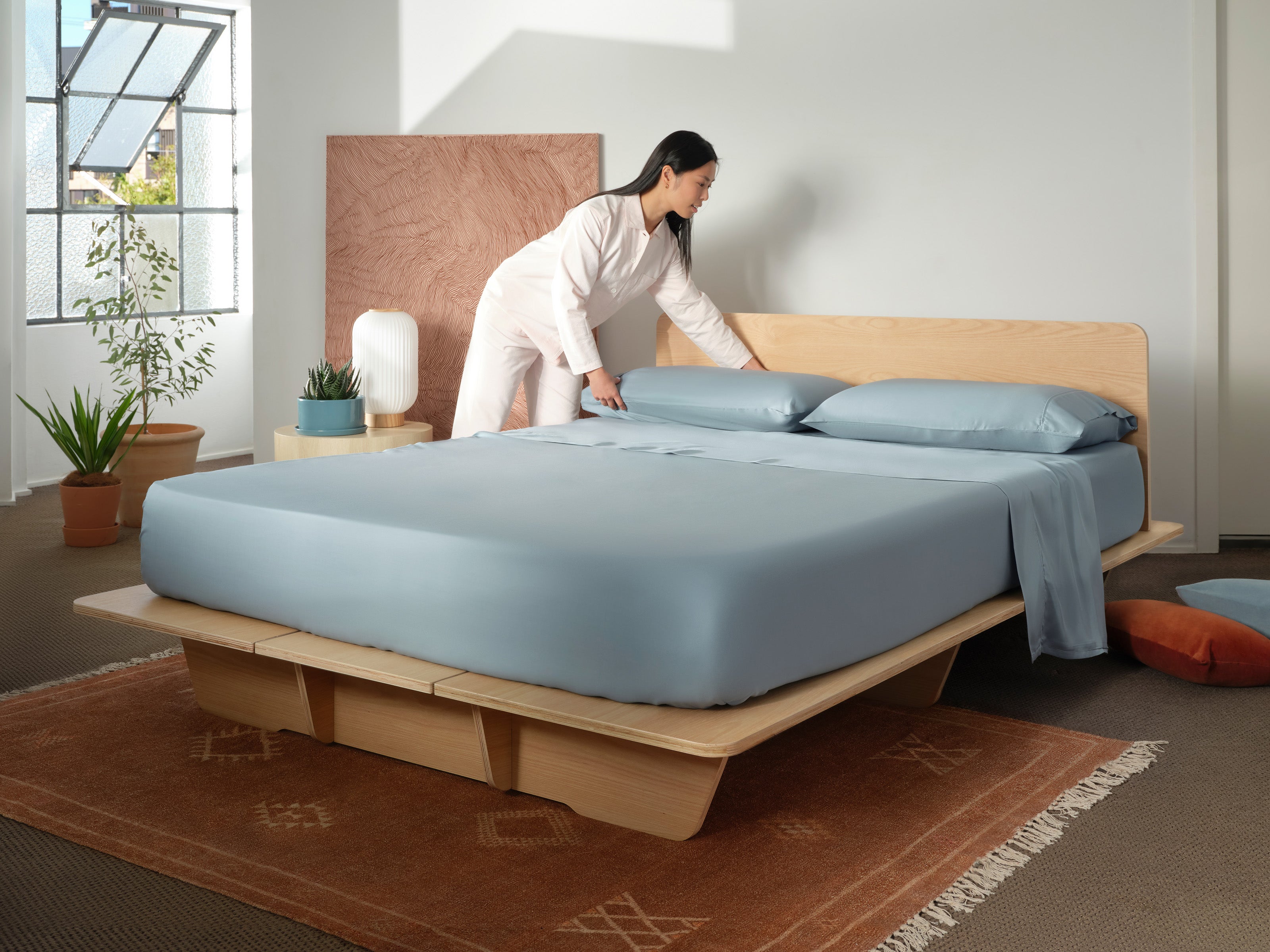 Woman making a bed with light blue sheets in a room featuring a wooden platform bed, potted plants, and a tan rug on a grey carpet.