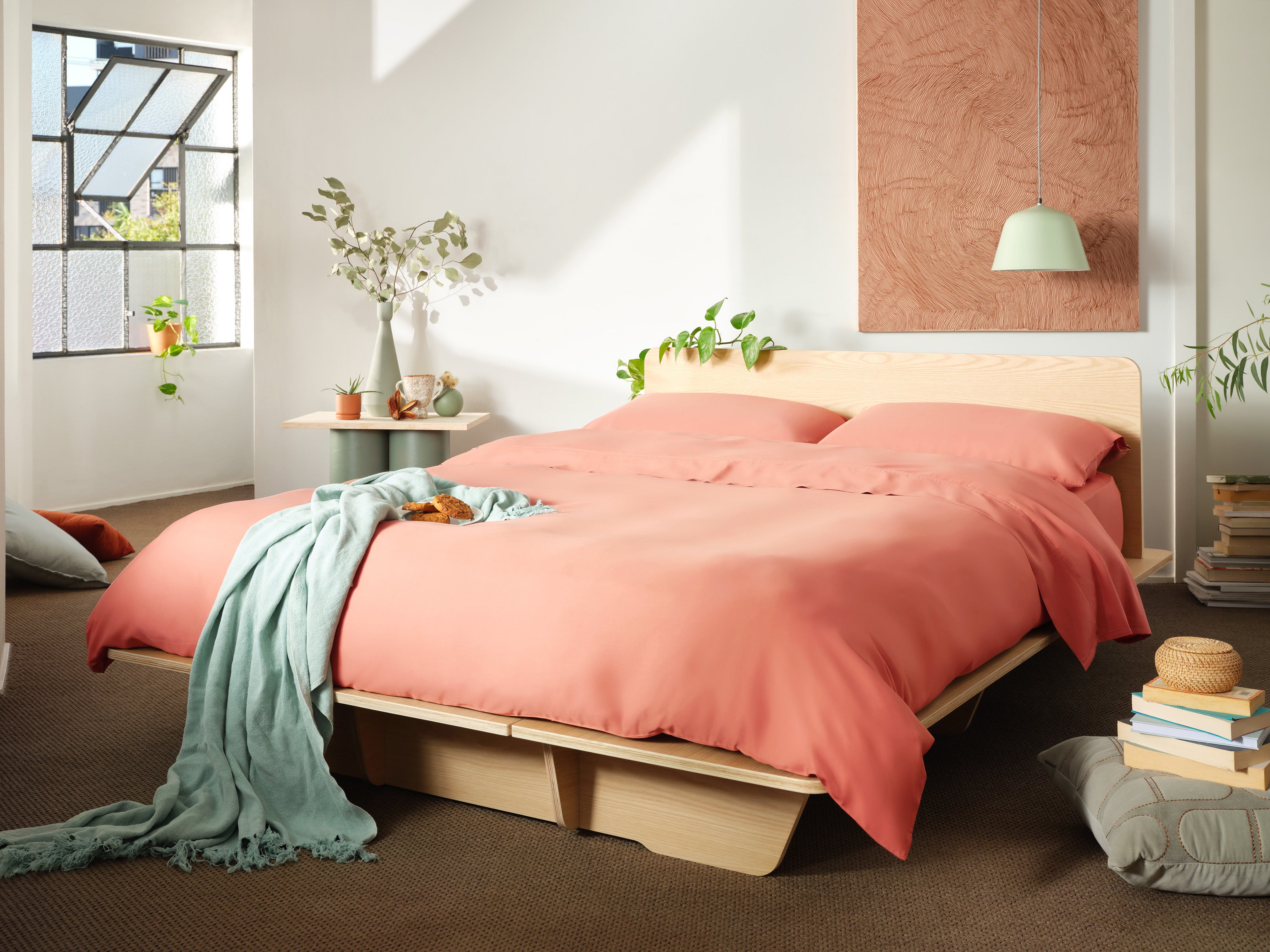 Modern bedroom with light wood bedframe, coral bedding, light teal throw, and decorative plants. Large window, pendant lamp, and books on floor.