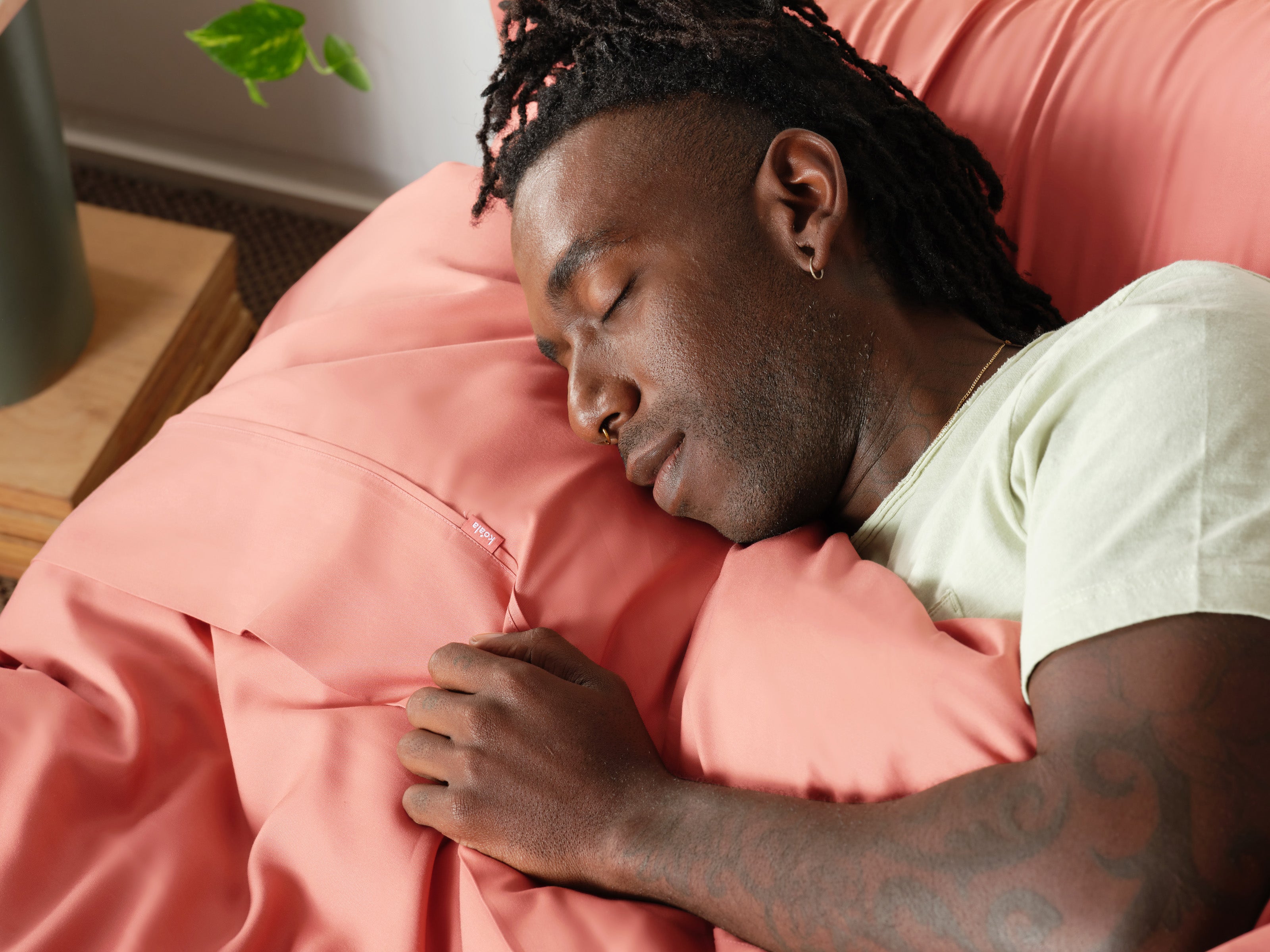 Man sleeping on a bed with soft pink sheets, wearing a light green shirt. A plant and wooden furniture are in the background.
