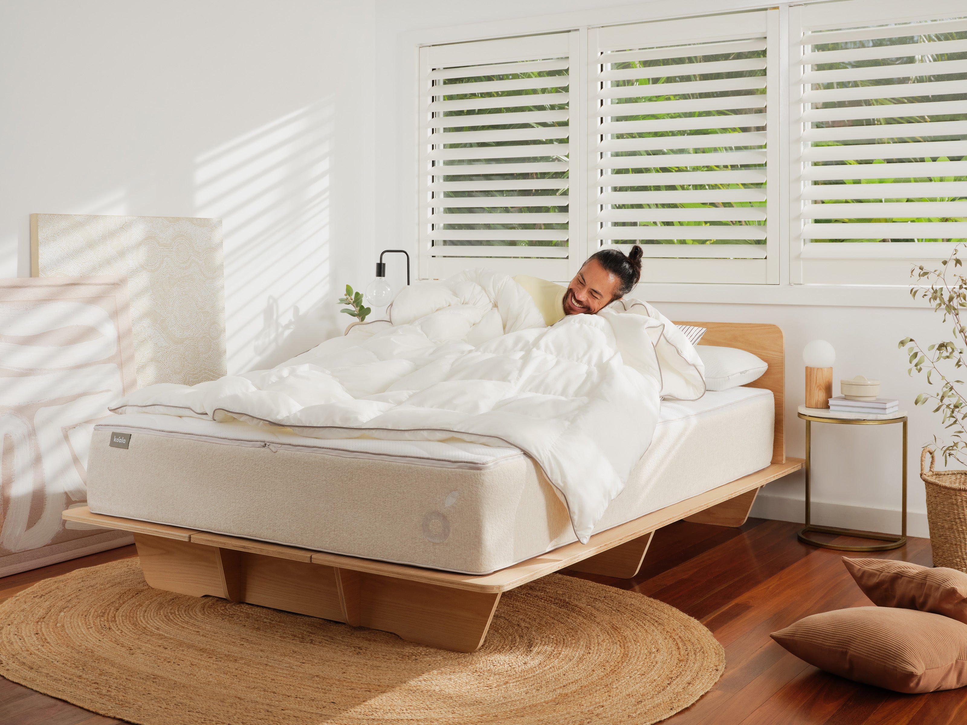 Man relaxing on a bed with a wooden frame, white bedding, in a cosy bedroom with natural light, wooden floors, and indoor plants.