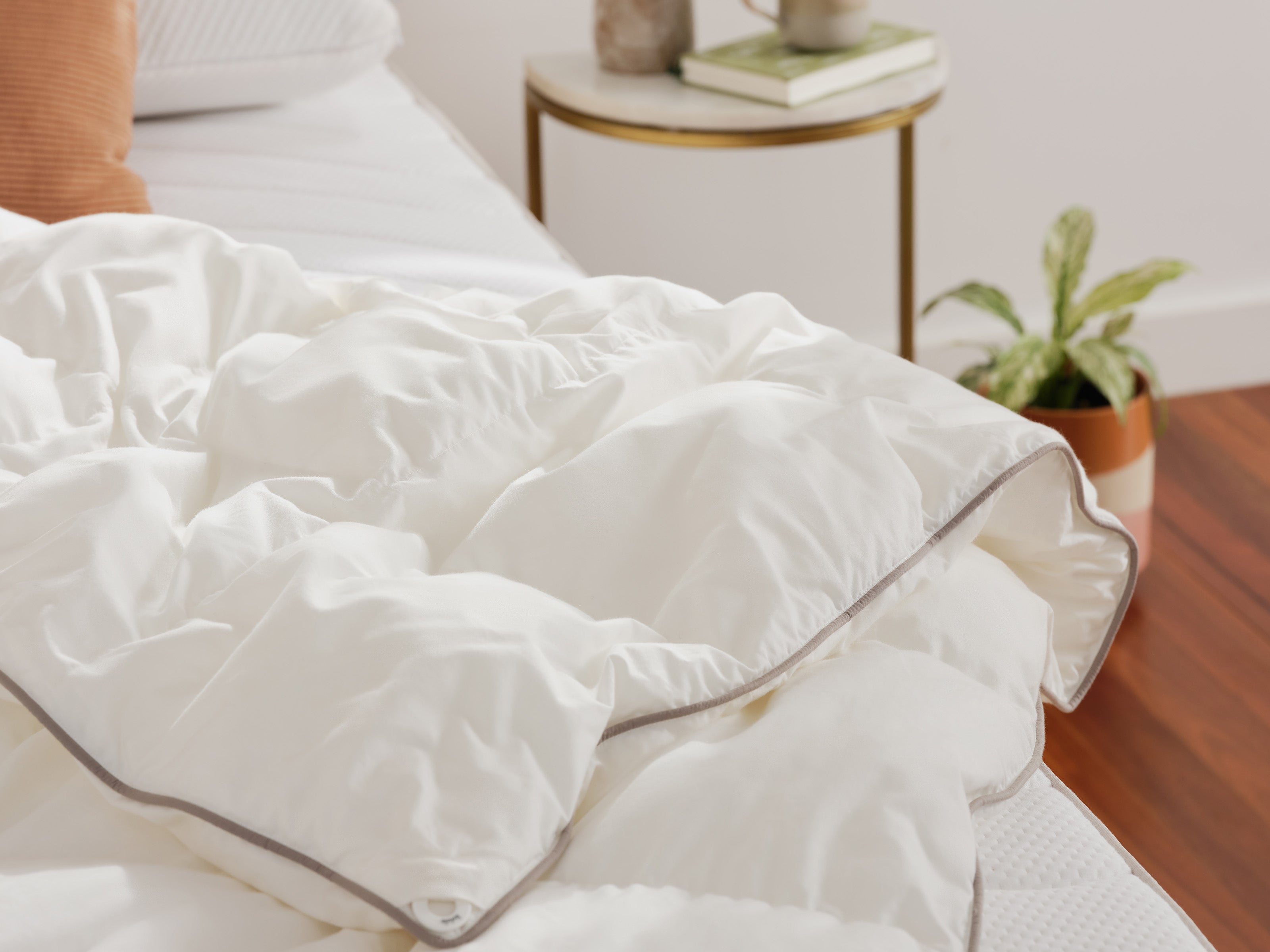 White quilt with grey trim on a bed, beside a gold-accented round side table with books and a plant in a pot on wooden floor.