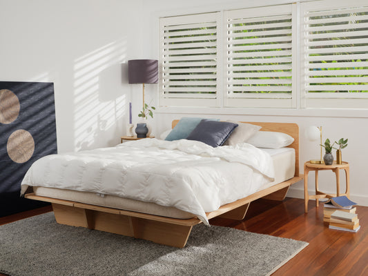 Modern bedroom with wooden bed frame, white quilt, grey carpet, and bedside tables. Large window with shutters; minimalist decor.