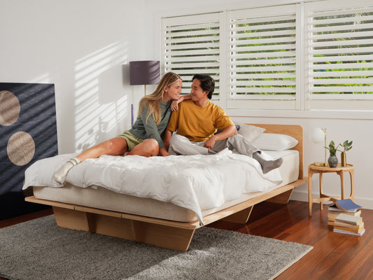 Couple sitting on a natural wood bed with white bedding in a modern bedroom. Features include a rug, painting, and large windows with shutters.