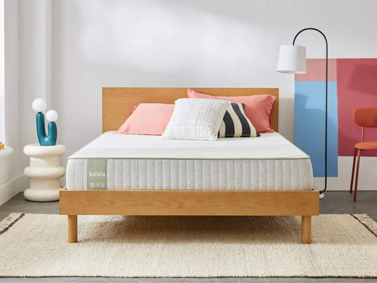 Minimalist bedroom with a light wood bed frame, white mattress, pink and striped cushions, modern lamp, and abstract wall art; cosy and stylish décor.