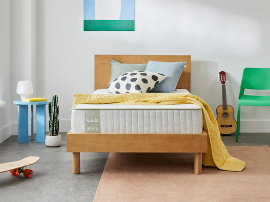 Modern bedroom with a wooden bed frame, light grey mattress, and patterned cushions. A green chair, guitar, and cactus accent the room.