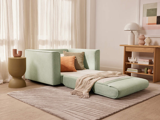 Pastel green modular sofa bed in a cosy living room with a light grey rug, wooden side table, and decorative accents.