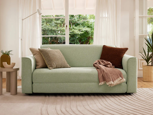 Light green sofa with beige and brown cushions, draped with a blanket. Surrounded by a floor lamp, plants, and large window view in a cosy living room.