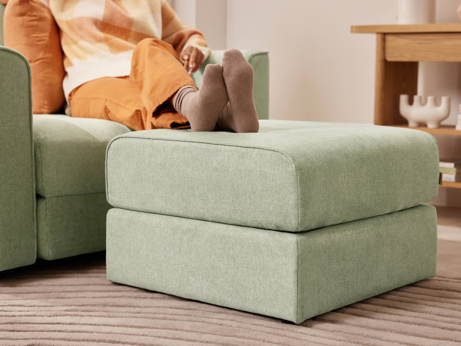 A person with crossed legs rests on a green ottoman next to a matching couch. The room has a beige rug and a wooden side table.