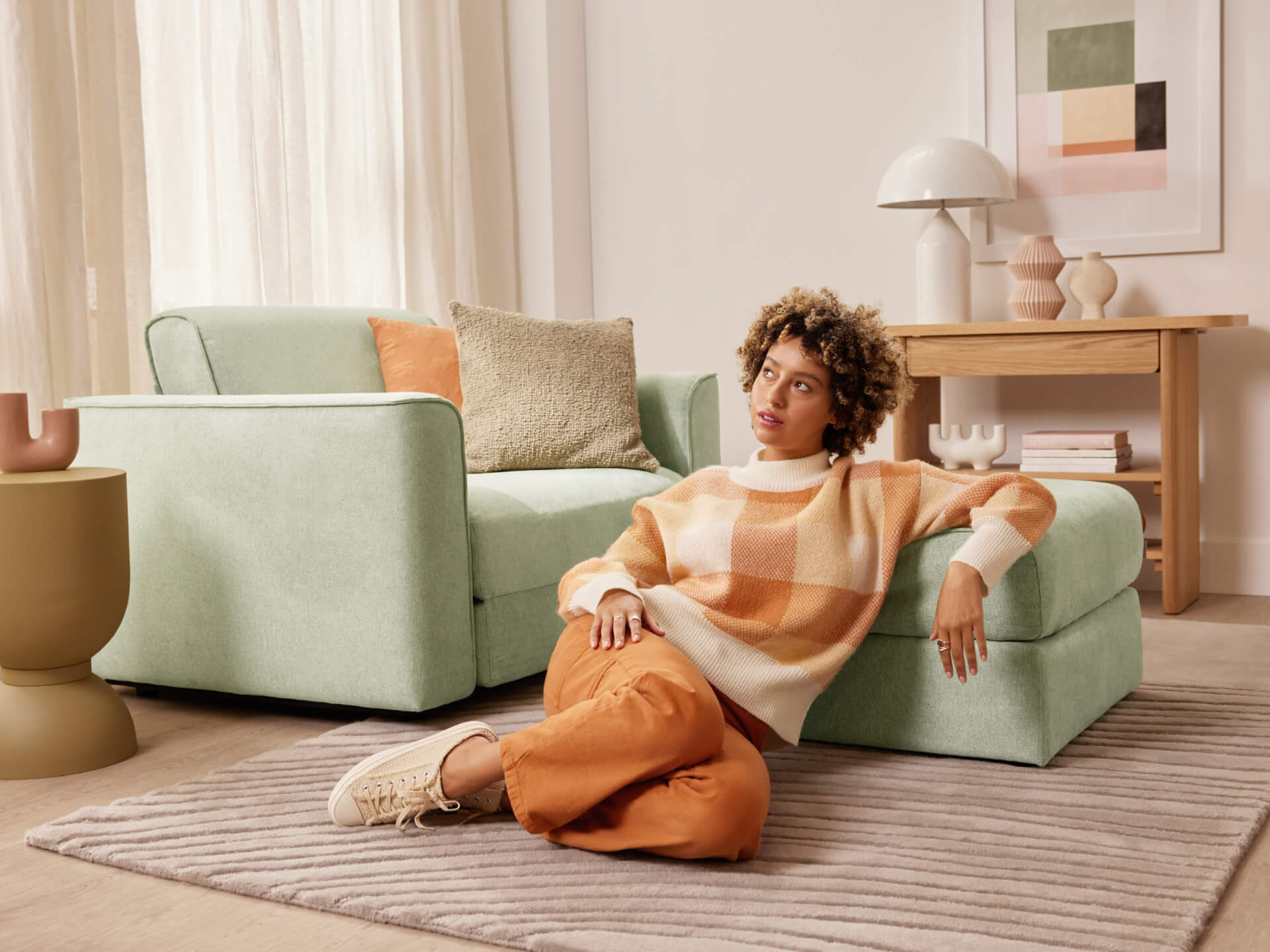 Woman sitting on a grey rug, leaning against a light green sofa with orange and beige cushions. A wooden table and artwork are in the background.