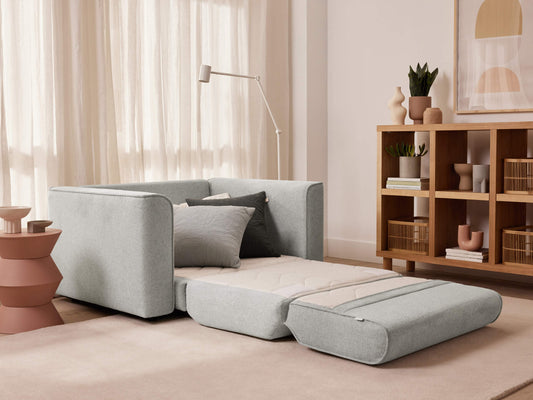 Light grey sofa bed extended in a modern living room with a wooden bookshelf, decorative items, and a floor lamp. Neutral-toned decor.