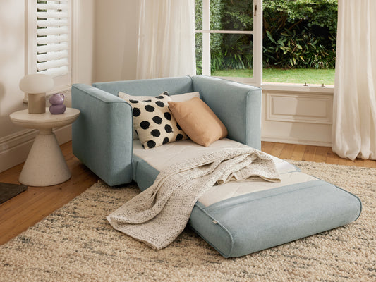 Light blue sofa bed with beige and black cushions and a cream throw, on a beige rug near a window. White side table with lamp on the side.