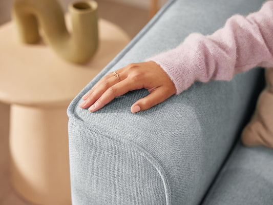 Hand resting on a light blue fabric couch arm, with a beige table and decorative vase in the background.