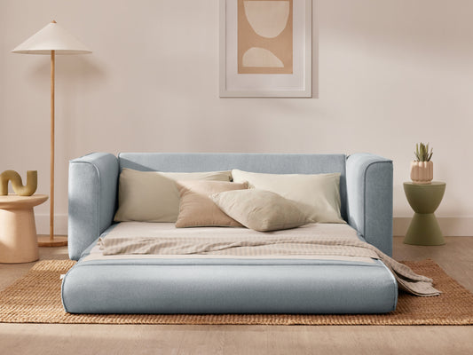 Light blue sofa bed with cream cushions and a beige throw on a woven rug. Minimalist decor includes a floor lamp and abstract wall art.