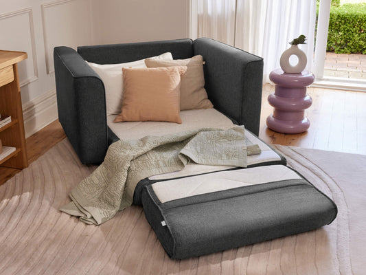 Dark grey armchair with beige cushions and a matching ottoman unfolded into a bed, placed in a cosy room with light wood flooring.