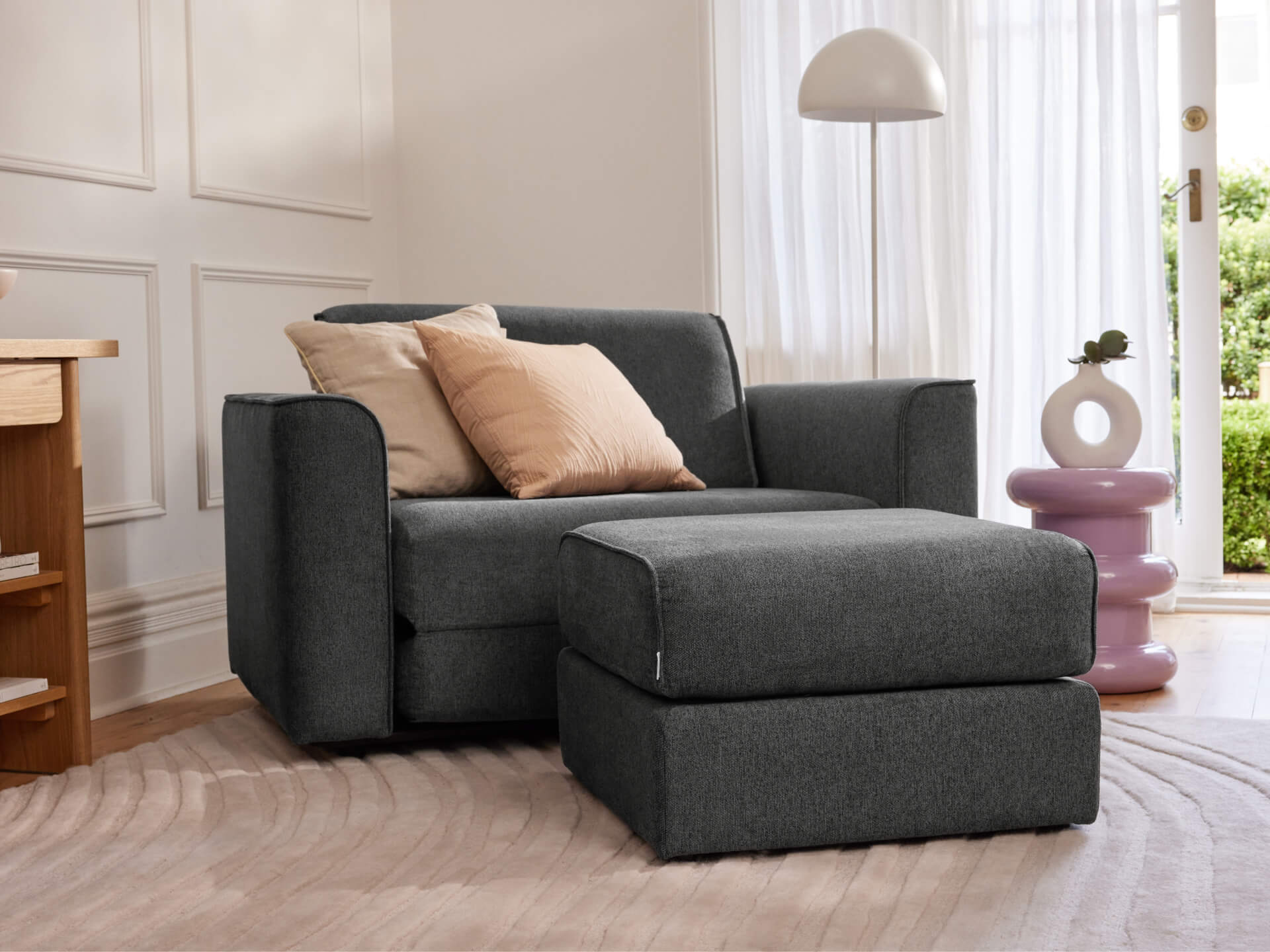 Dark grey armchair and ottoman with beige cushions in bright room; modern decor includes a round lamp and lavender side table.