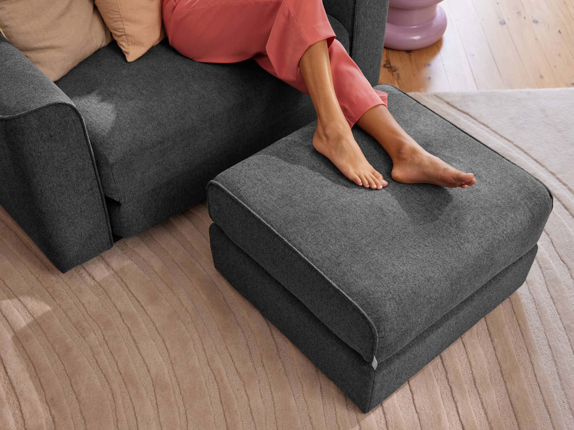 Grey sofa and ottoman with a person lounging on it, pink pants, beige cushion, on a light brown rug and wooden floor.