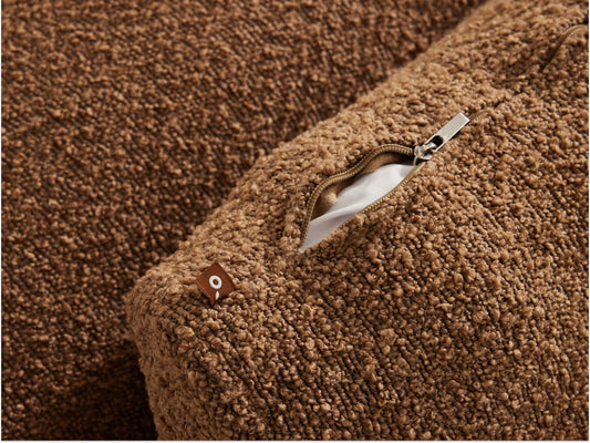 Close-up of a textured tan cushion with a visible zip and white lining. A small brown tag with a white logo is seen beside the zip.