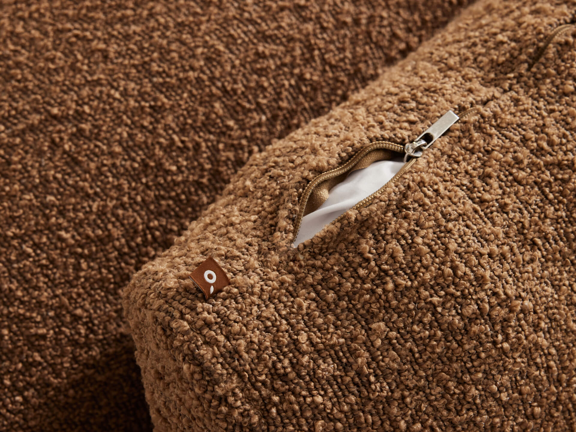 Close-up of a brown textured cushion cover with an open metal zipper, revealing white fabric inside. A small label is visible near the zipper.