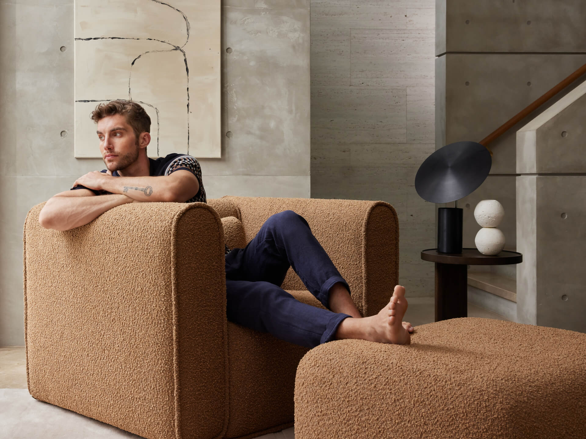 Man lounging on a tan armchair with a matching ottoman in a modern room. Grey concrete walls, abstract art, and a modern lamp are visible.
