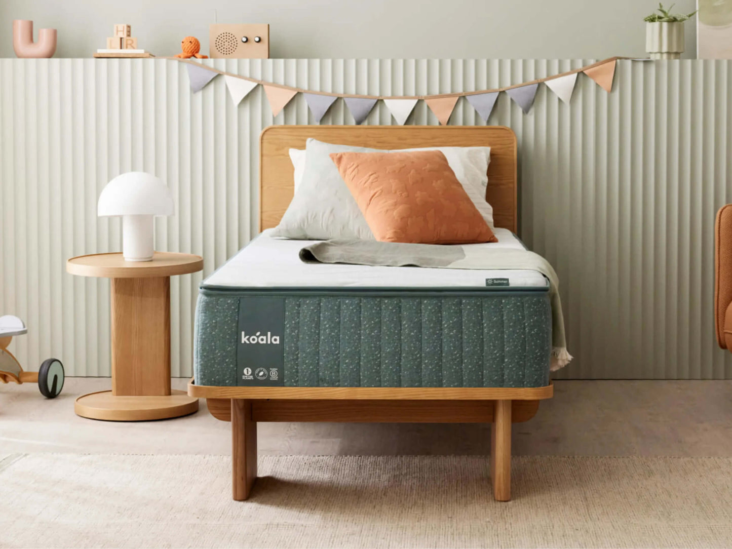 A bedroom with a wooden bed featuring a grey mattress and peach cushion. A round wooden side table with a white lamp is nearby. Decorative bunting hangs above.