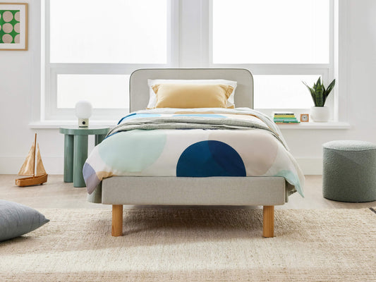Modern bedroom with a single bed, abstract circle duvet, light grey headboard, and wooden legs. White walls, large windows, green side table.