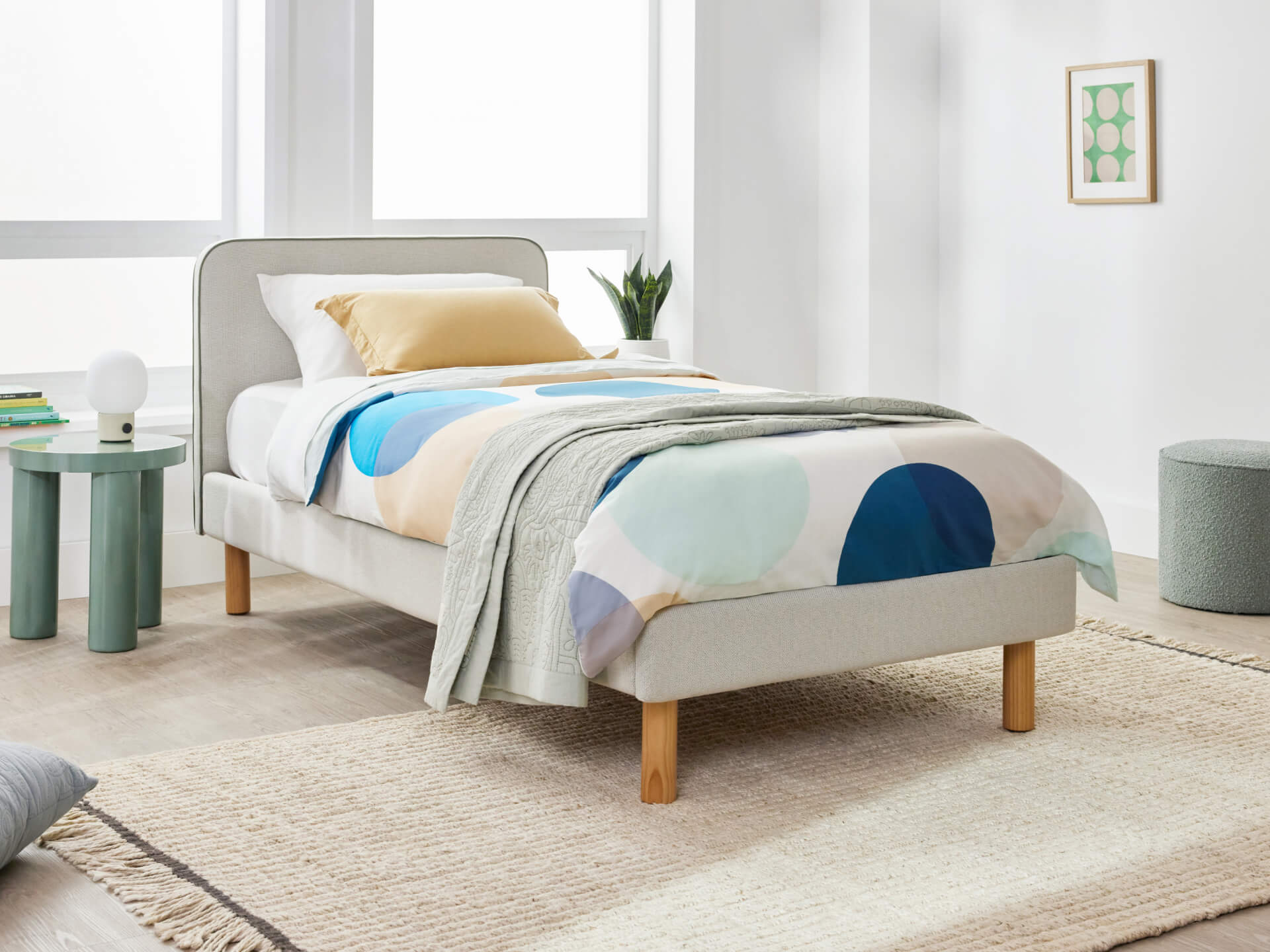Stylish bedroom with a single bed featuring light grey upholstery and wooden legs. Colourful duvet, sage side table, green plant, and framed art.
