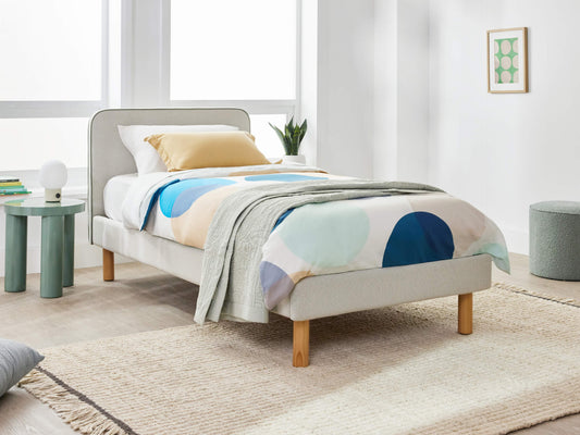 Stylish bedroom with a single bed featuring light grey upholstery and wooden legs. Colourful duvet, sage side table, green plant, and framed art.