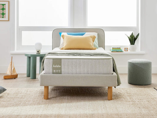 Minimalist bedroom with a single bed featuring light grey upholstered frame, yellow and blue cushions. Green side table, potted plant, and round pouf.