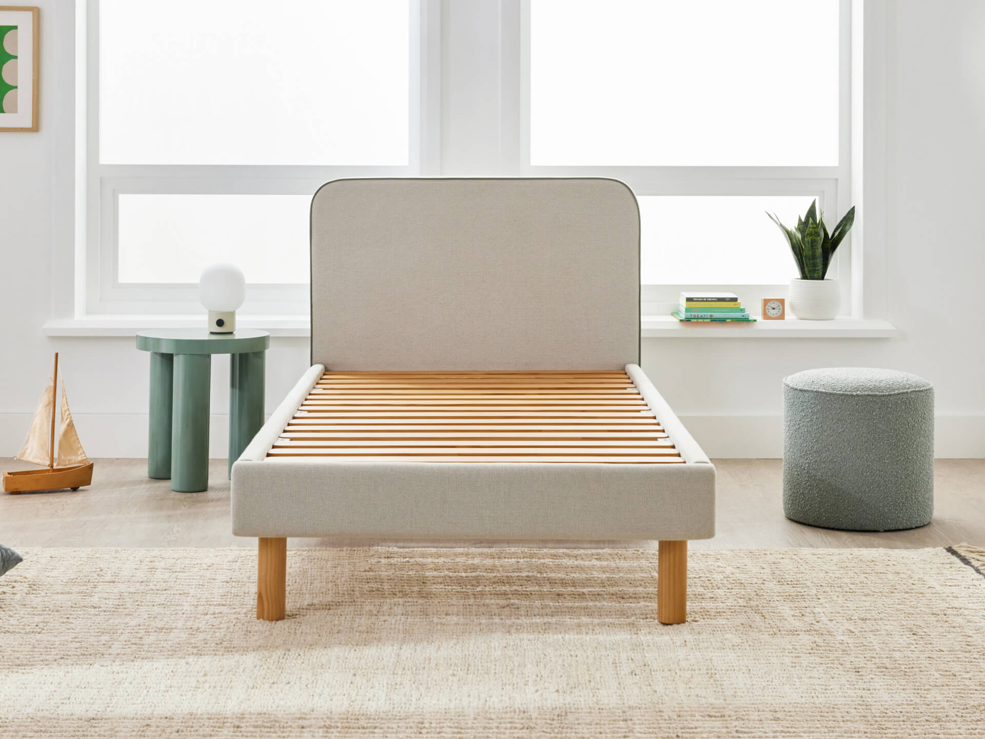 Light grey bed frame with wooden slats in a bright room. Nearby, there is a small green side table, a grey ottoman, and potted plants.
