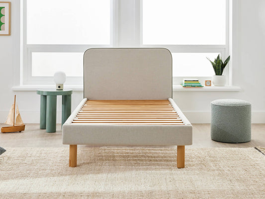 Light grey bed frame with wooden slats in a bright room. Nearby, there is a small green side table, a grey ottoman, and potted plants.