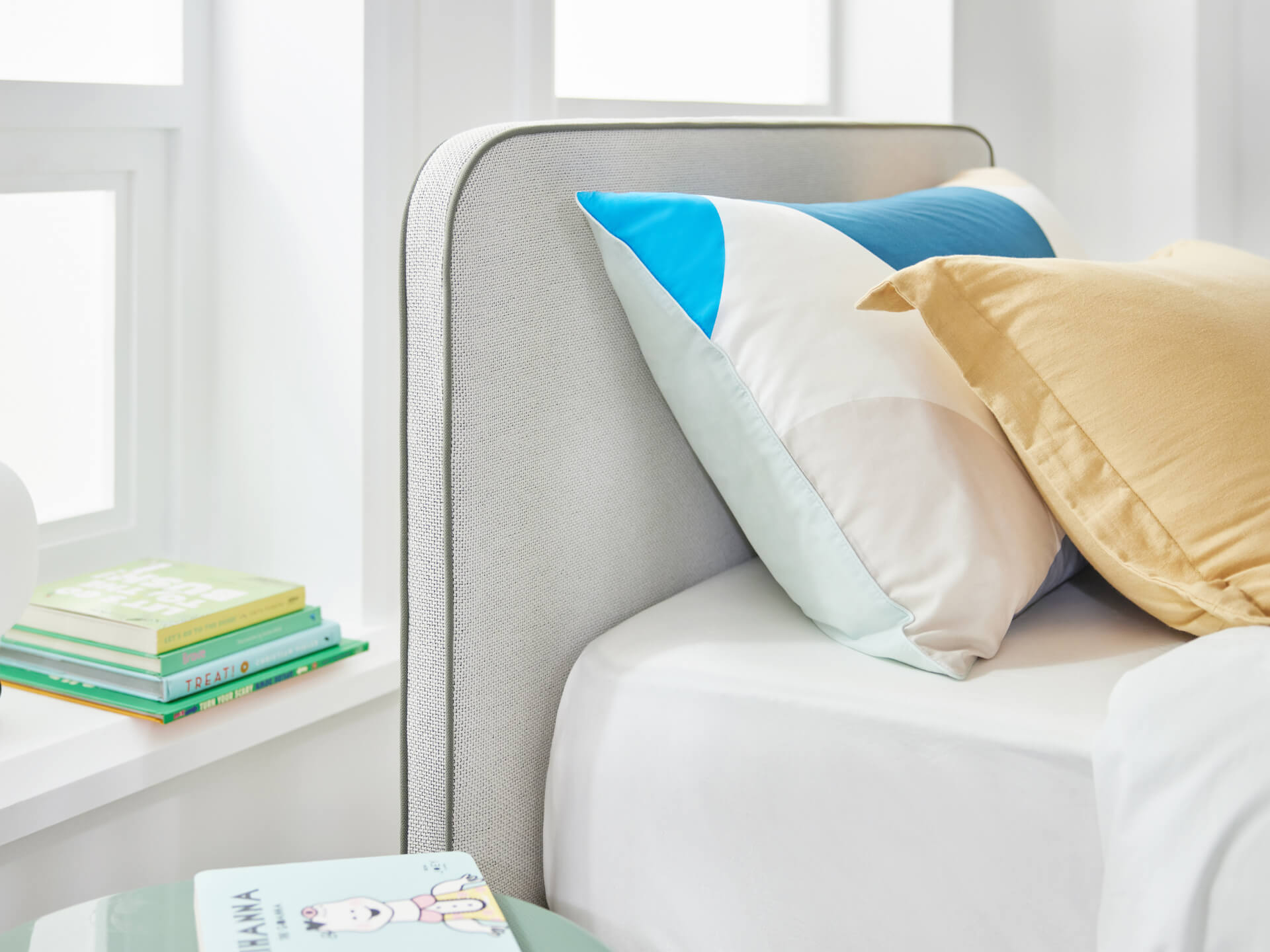 Minimalist bedroom with a light grey upholstered headboard, colourful pillows, and a stack of books on a white windowsill.