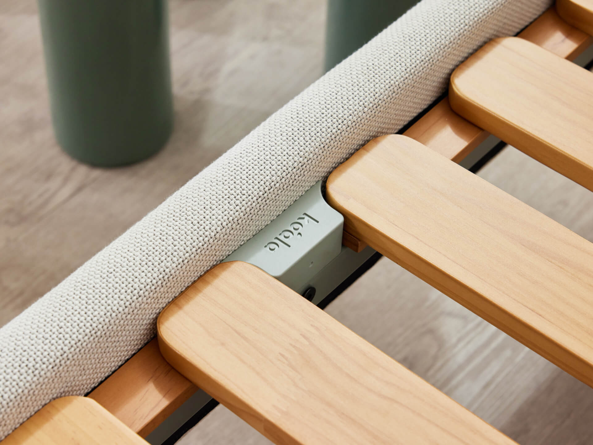 Close-up of a light wood bed slat frame with a beige fabric edge and a metal connector, set against a light wood floor.