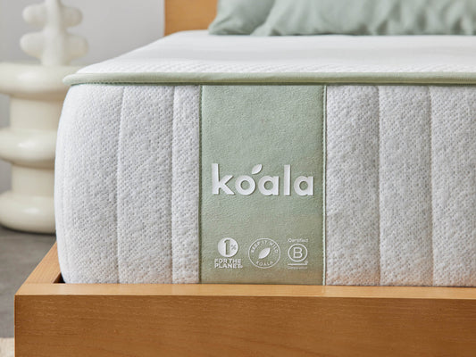 Close-up of a mattress corner with a light grey quilted fabric and green brand label. Set on a wooden bed frame. White and green bedding visible.