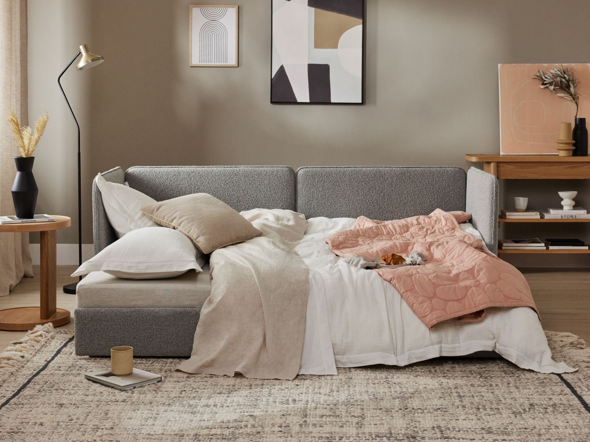 Cosy living room with a grey sofa bed, covered in beige and pink bedding. Framed art, floor lamp, and decor on a wooden shelf complete the scene.
