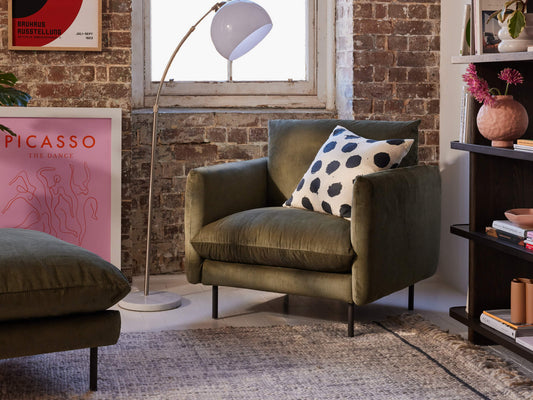 Olive green armchair with black polka dot cushion, against an exposed brick wall, next to a white floor lamp and decorative items.