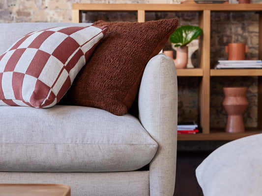 Light grey sofa with checkered and brown cushions, against a brick wall. Wooden shelf with books and plants in the background.
